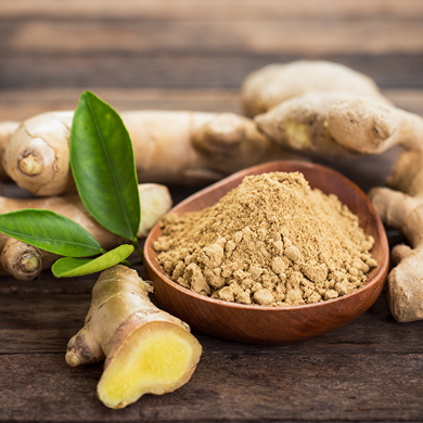 Fresh ginger roots and green leaves surrounding a wooden bowl of ground ginger powder